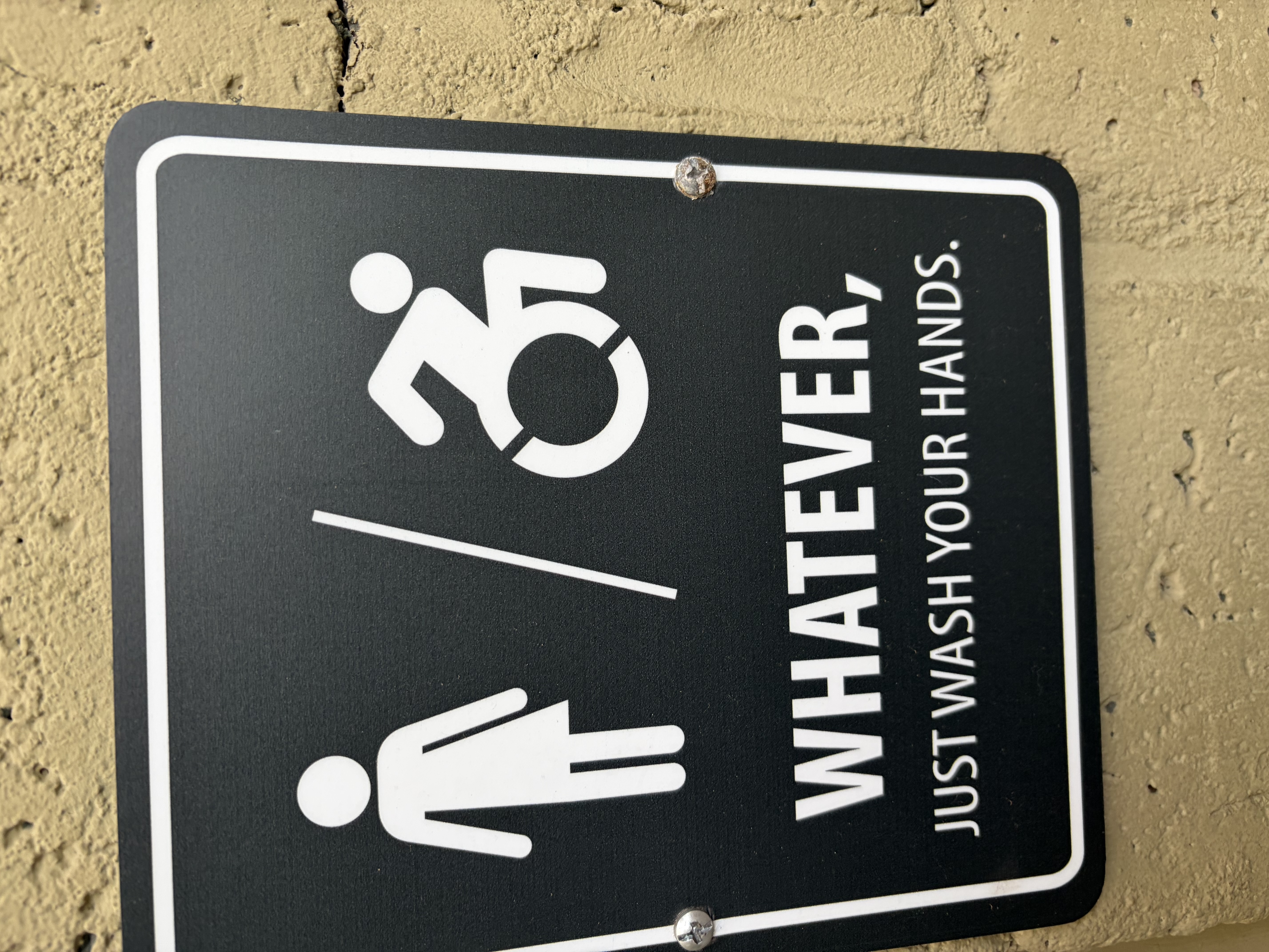 A restroom sign with two figures reading: WHATEVER, JUST WASH YOUR HANDS.