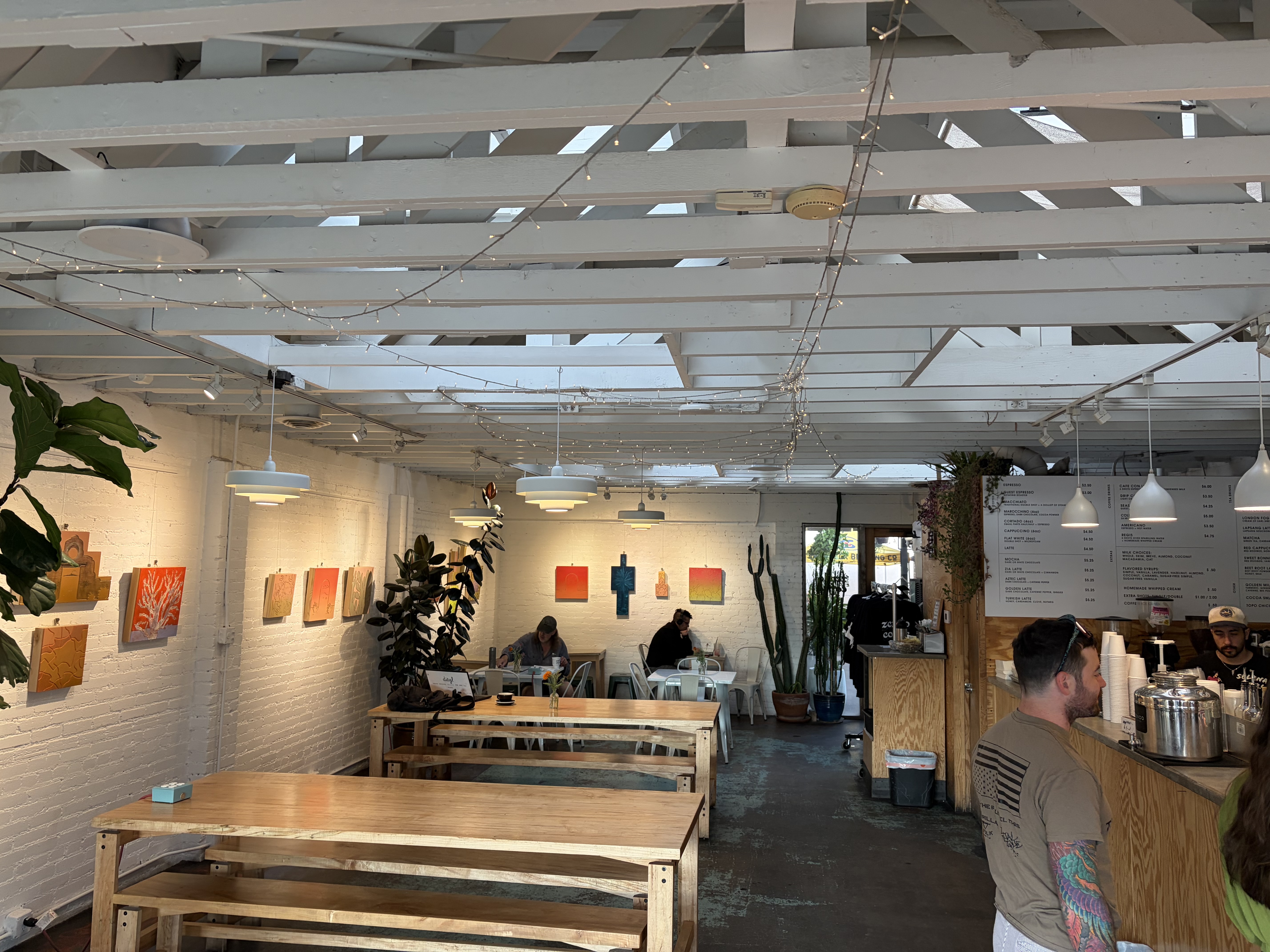 Interior: white painted brick, exposed white roof joists, string lights, long communal wooden tables, local paintings, menu boards behind a wooden counter.